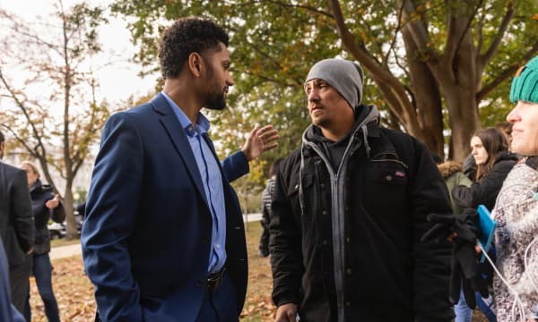 A dark skinned man with a beard wears a navy suit and stands sideways, one arm outstretched to a white man wearing a gray beanie and a black winter coat.