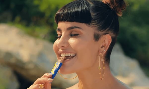 Girl with dark hair in a bun and long gold earrings smiling with her eyes closed as she eats a chocolate bar
