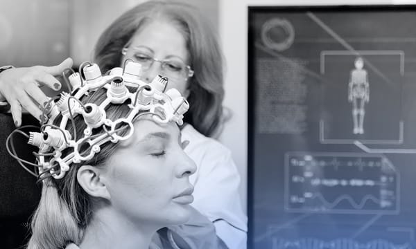 A woman in a lab coat wearing glasses holds a device over another woman's head in a black-and-white photo.
