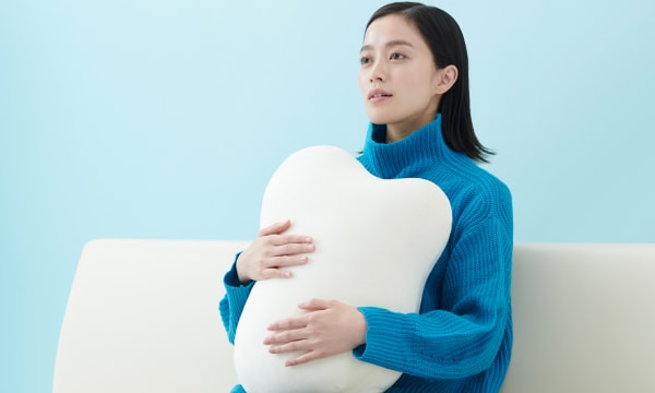 A woman with light skin and dark hair holds a pillow to her chest and looks to the left of the frame.