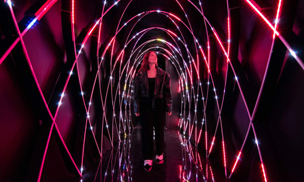 A woman with light skin and long dark hair walks towards the viewer through a magenta, lit-up tunnel, and looks up and to the viewer&#039;s right.