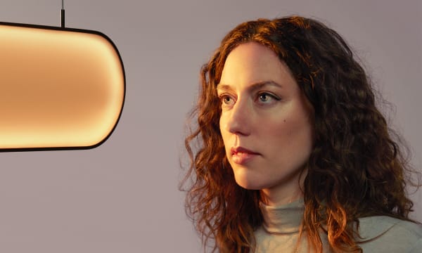 A woman with light skin and curly, long auburn hair is staring to the left at a pill-shaped light fixture.