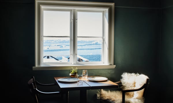 A table is set in front of a window in a room with dark green walls and two chairs. Through the window is an arctic ocean scene.