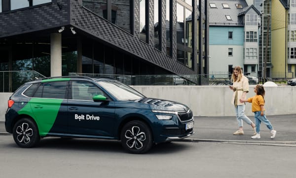 A woman with blonde hair holds a young girl's hand and looks down at a cell phone. They are walking towards a car that reads "Bolt Drive"