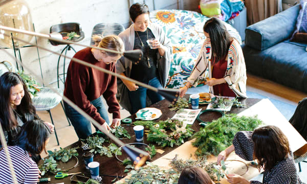 A group of seven people are gathered around a table working with greenery and twine.