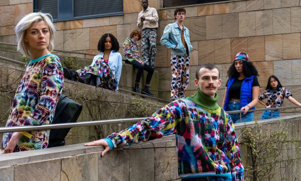Models stand in an urban space with stone walls wearing similar, colorful animal-print patterned sweaters. A man and a woman stand in the forefront, and 6 models stand behind them on steps.