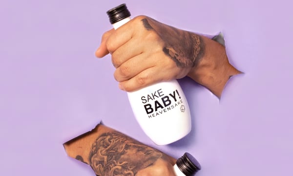 A tan hand with tattoos reaches through a purple backdrop and clutches a white bottle that reads &quot;SAKE BABY! HEAVENSAKE&quot;