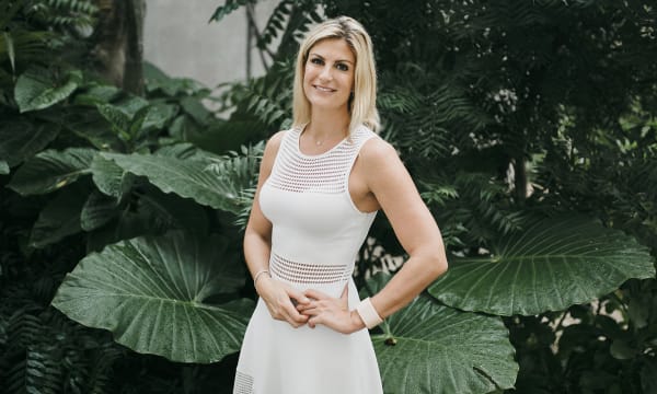 A white woman with shoulder-length blonde hair stands in front of a leafy green wall and smiles at the camera with one hand on her hip, wearing a white dress.