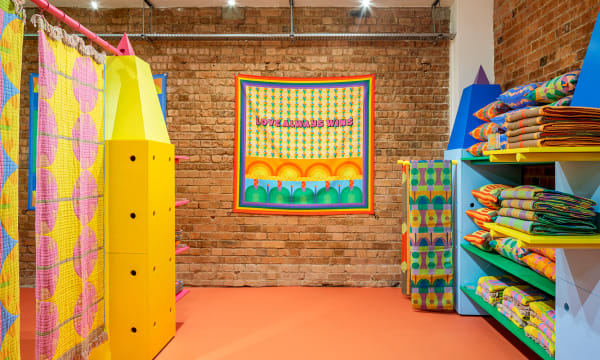 A bright retail space, with yellow shelving and quilted tapestries on the left, green and blue shelving on the right, orange flooring, and a bring wall with a tapestry hanging in the back that reads &quot;Love always wins&quot;