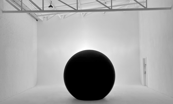 A large black cylinder a white room, with a light subtly shining from behind it and some scaffolding above it.