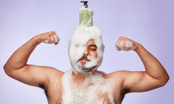 A white bare chested man with a beard strikes a tough guy pose against a blue background his head is covered in foam and a bottle branded Stuff is perched on his head