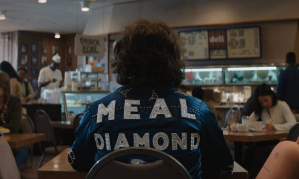 Image of a person sitting at a table in a diner wearing a jean jacket that has the words Meal Diamond sequined on it. There's a salami with a face drawn on it in the chair next to him.