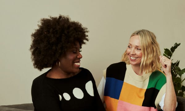 Two women chat on a sofa. One woman is black with an Afro hairstyle wearing a black sweater with white spots, and the other is a white woman with a wavy bob in a geometric colour block sweater.