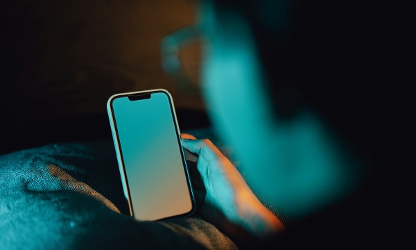 A dark image illuminated only by a blue-orange ombre emanating from a mobile phone that is just off the centre of the image, illuminating the blurred face of the person holding and looking at the phone.