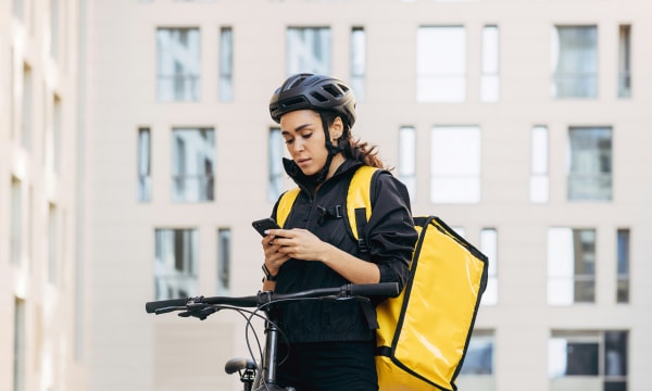 Delivery rider standing in front of a building with their bike and their yellow delivery rucksack, looking at their phone.