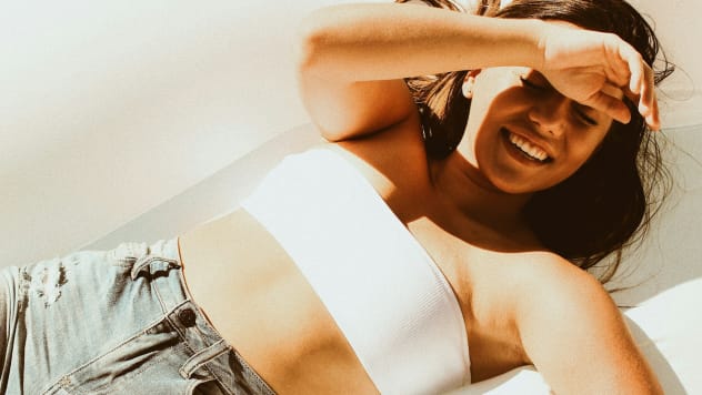 High-angle shot of a young woman lying down outdoors in intense sunlight, shielding her eyes with her arm while tanning in a white top and denim shorts.