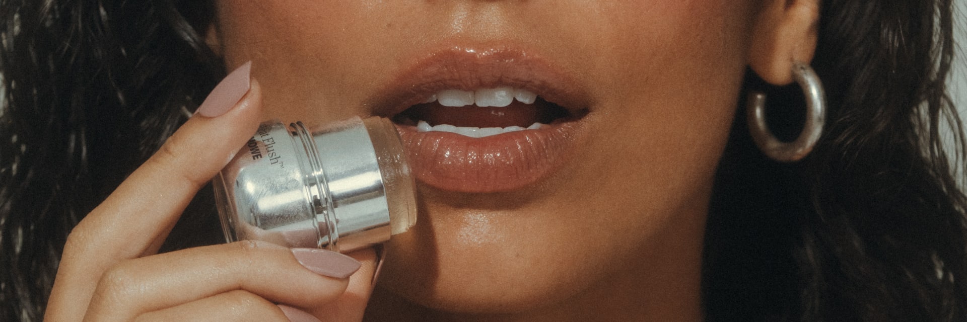 A tanned brown haired fresh-faced woman with her eyes closed holds a jar with a silver lid close to her lips which are slightly parted.