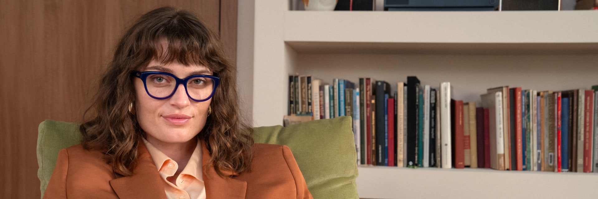 A woman wearing bold blue glasses and a brown blazer sits confidently in a cozy, book-filled room.