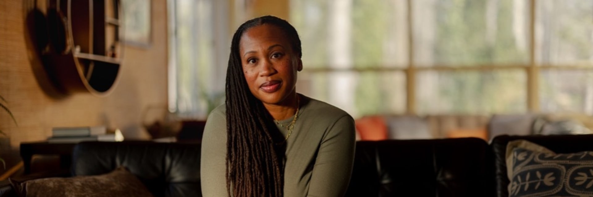 A woman with long braided hair sits on a black leather couch in a warmly lit living room. She is wearing a green sweater and blue pants, with her hands clasped together in front of her. The background features large windows with natural light streaming in, showcasing trees outside.