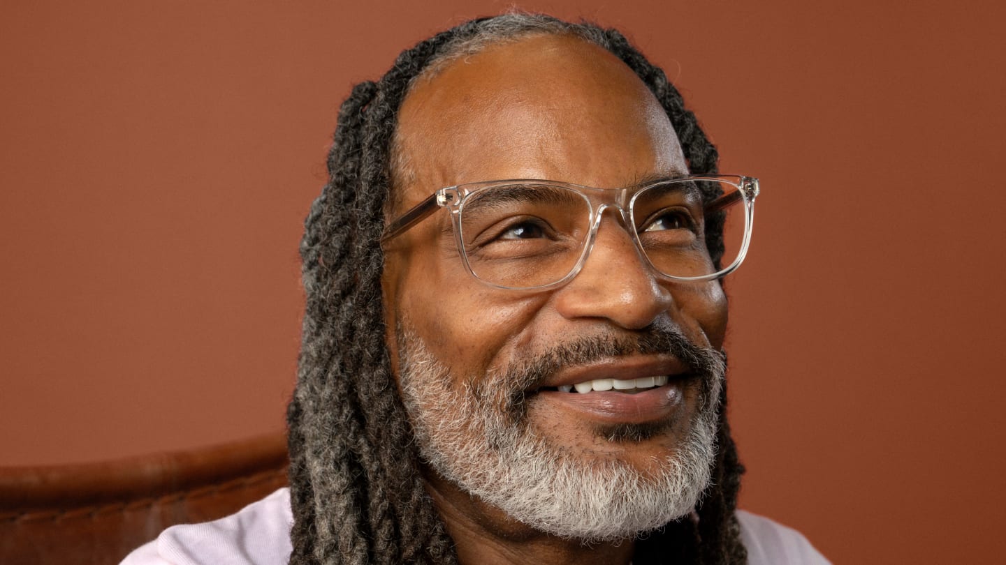 A smiling man with gray hair and clear glasses looks upward, set against a warm rust-colored background.
