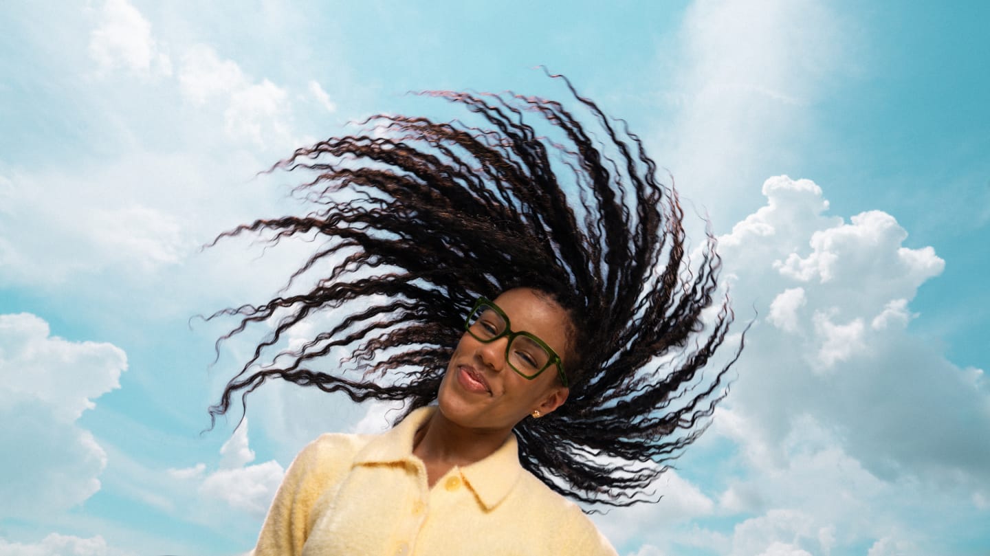 A smiling woman with flowing hair wears green glasses, set against a bright blue sky with clouds.