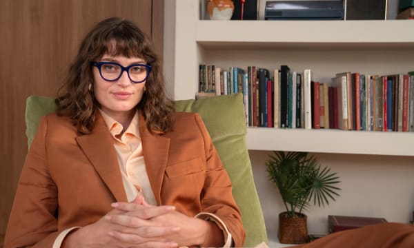 A woman wearing bold blue glasses and a brown blazer sits confidently in a cozy, book-filled room.