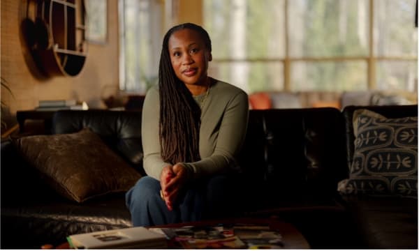 A woman with long braided hair sits on a black leather couch in a warmly lit living room. She is wearing a green sweater and blue pants, with her hands clasped together in front of her. The background features large windows with natural light streaming in, showcasing trees outside.