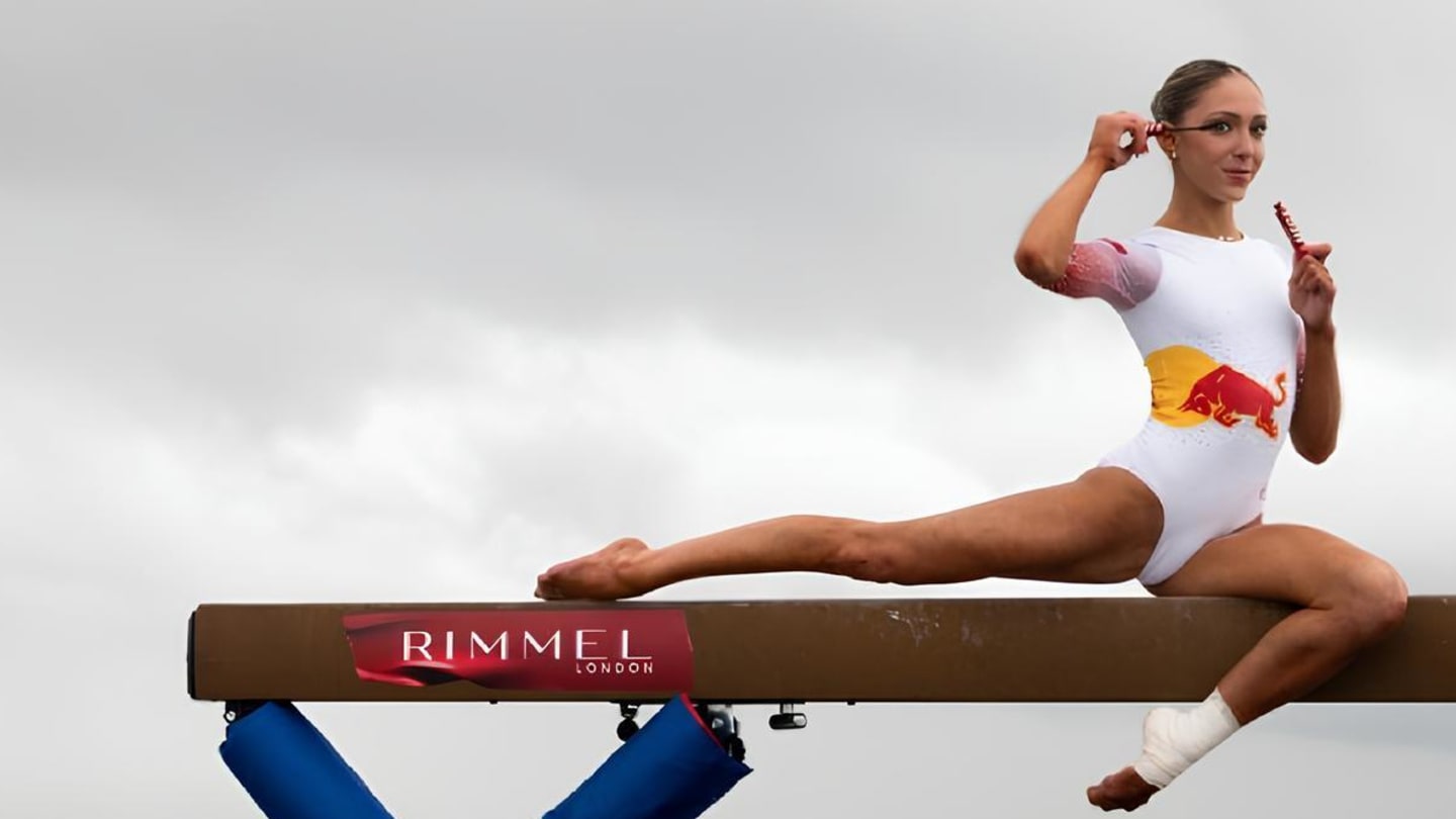 A female gymnast - Lily Smith - in a white leotard sitting in a split on an outdoor balance beam against an overcast city skyline background. She holds makeup products, applying them while balancing on the beam, which displays the Rimmel logo.