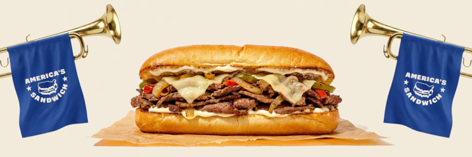 A close-up of a Philly Cheesesteak sandwich on parchment paper. The sandwich is flanked by two golden trumpets adorned with blue banners that read "America's Sandwich" with a sandwich icon. The background is a light beige color.