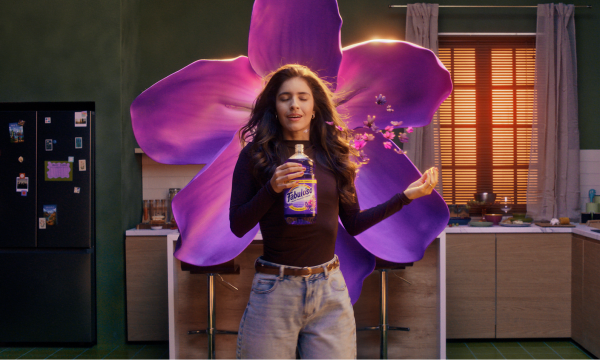 A woman stands in a vibrant kitchen holding a bottle of Fabuloso cleaner. Behind her is a large, glowing purple flower, symbolizing the product’s bold and transformative scent.