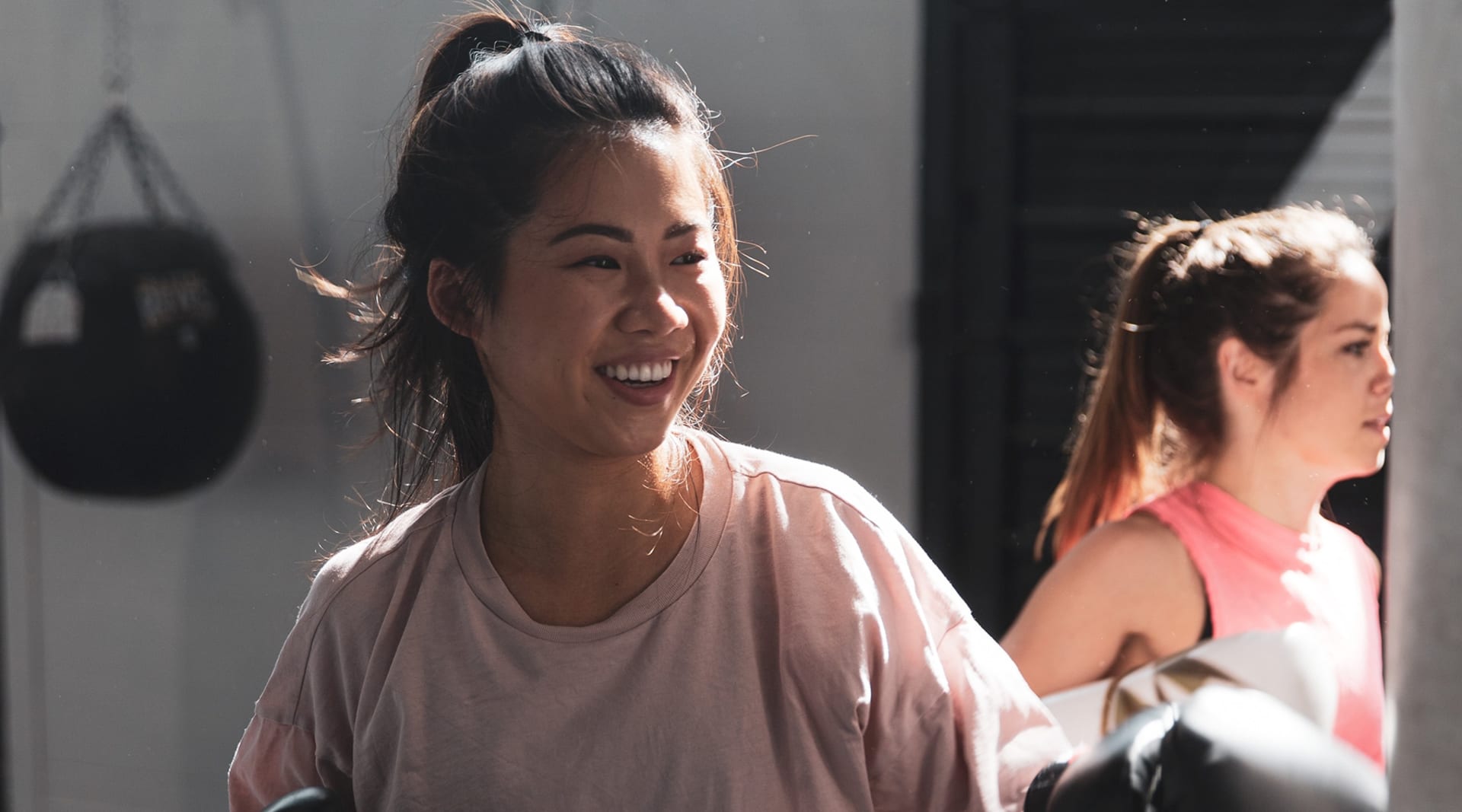 A woman with light skin and dark hair smiles as she works out. Her hair is in a high pony tail and she wears a pink T-shirt.