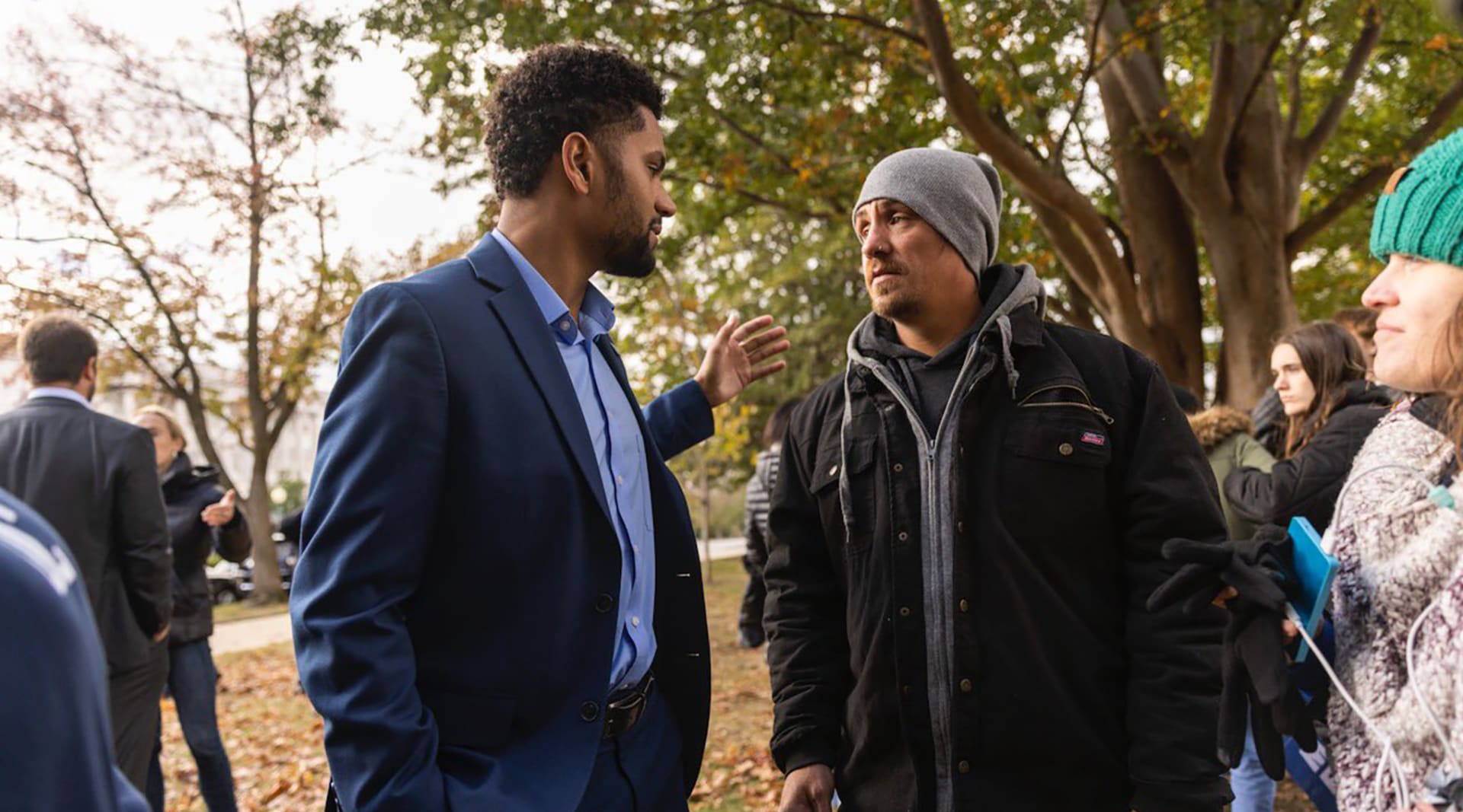 A dark skinned man with a beard wears a navy suit and stands sideways, one arm outstretched to a white man wearing a gray beanie and a black winter coat.