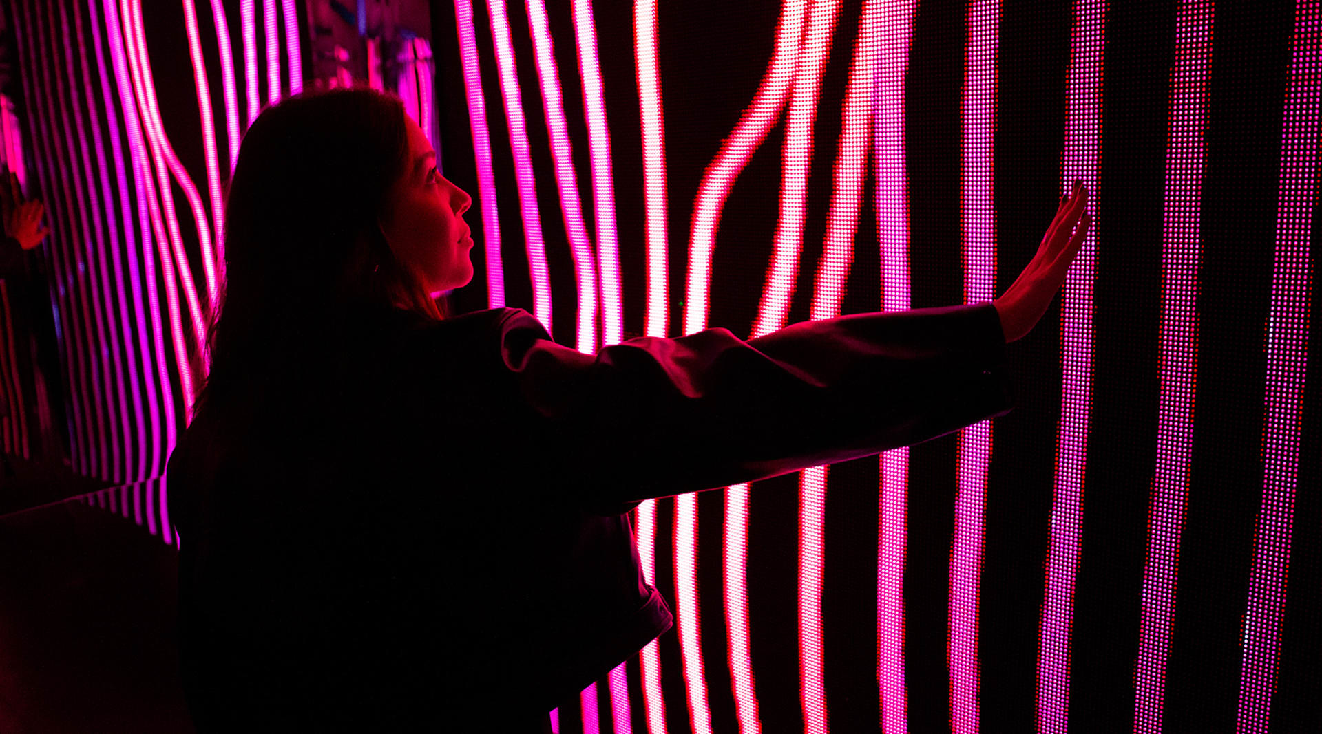 A woman with long hair stands in a magenta room with neon striped walls, her arm stretched out in front of her.