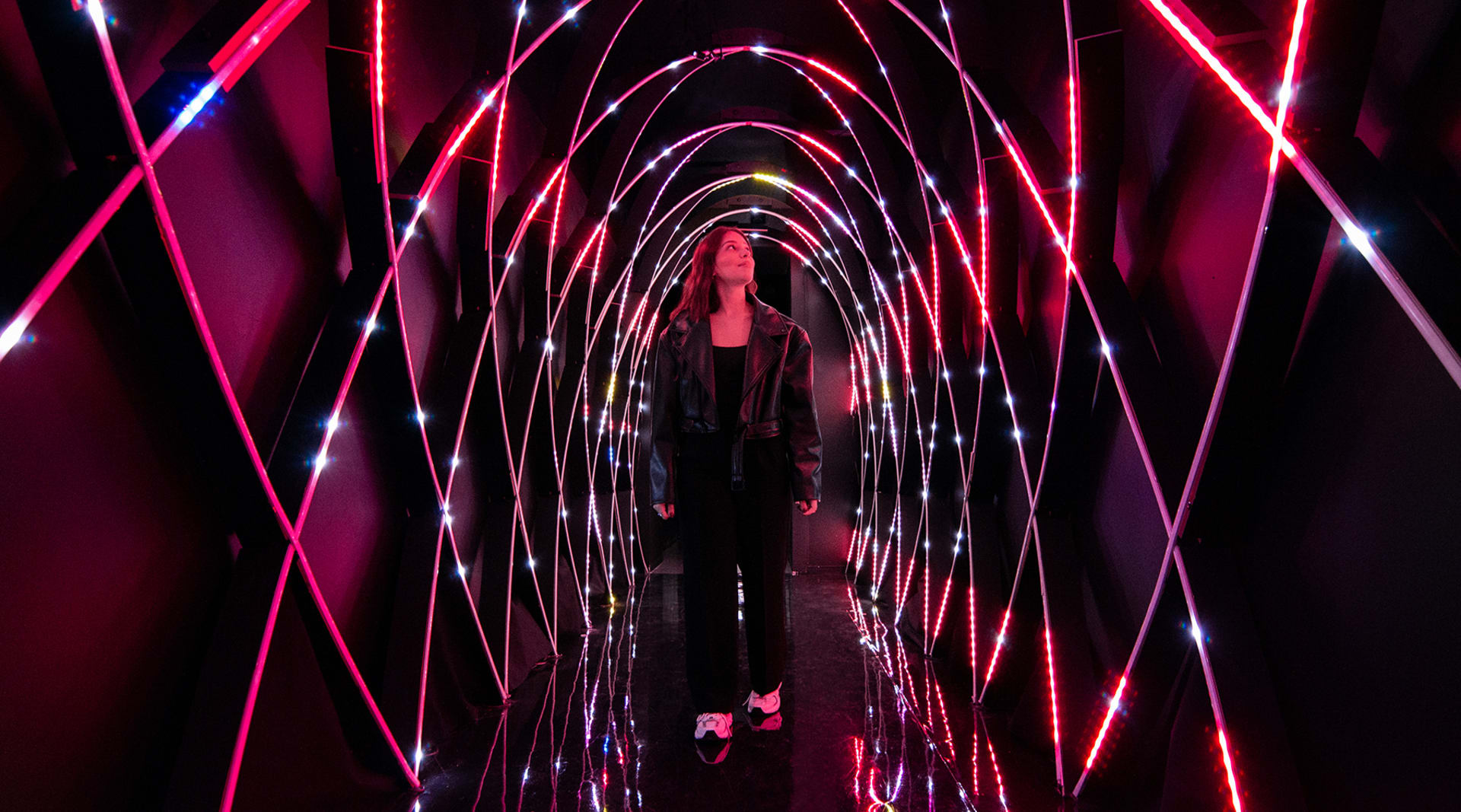 A woman with light skin and long dark hair walks towards the viewer through a magenta, lit-up tunnel, and looks up and to the viewer's right.