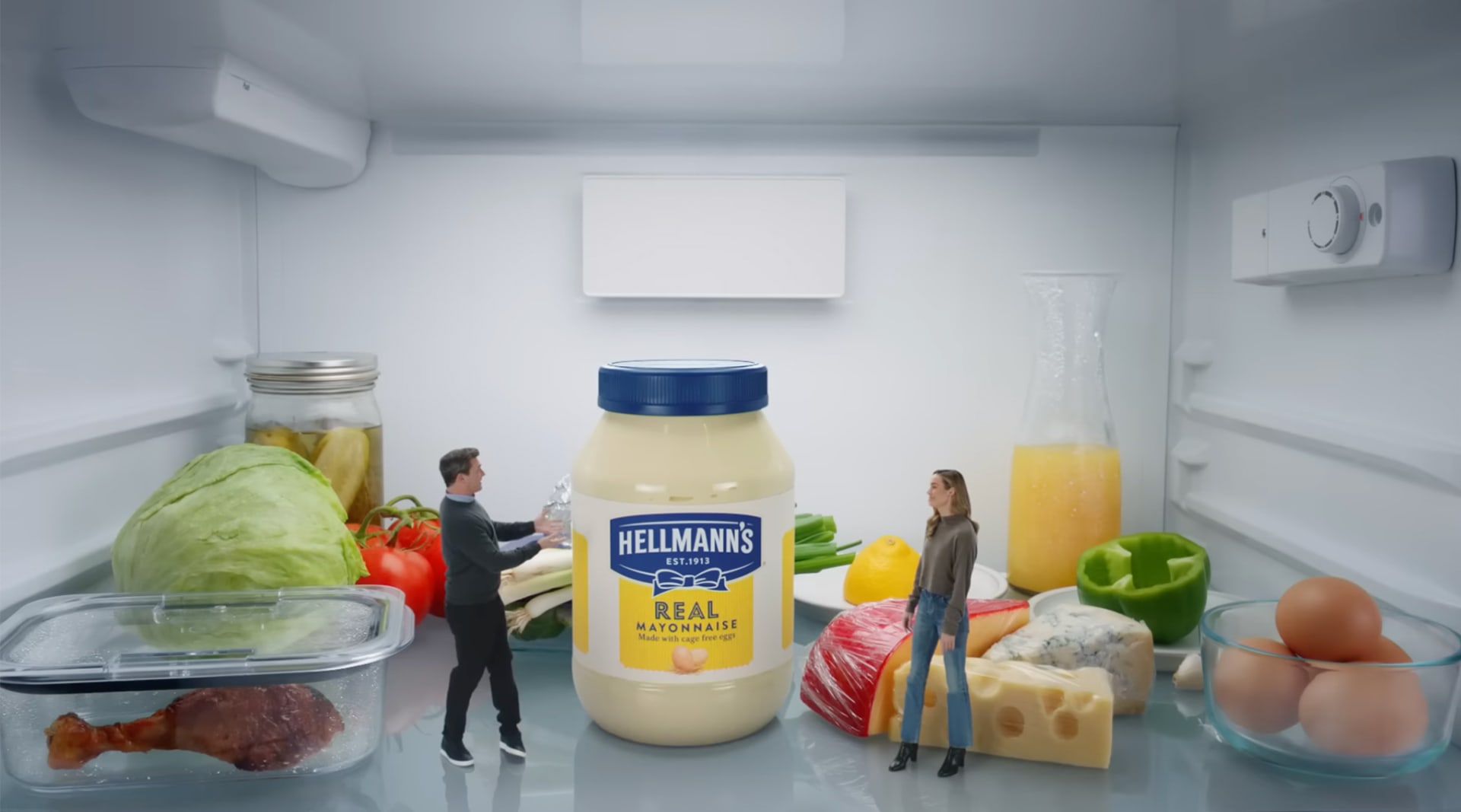 A man with dark hair and light skin stands opposite a woman with blonde hair and light skin. They are miniaturized and standing on a refrigerator shelf next to various produce items: an old chicken wing, lettuce, tomatoes, cheese, orange juice, eggs.