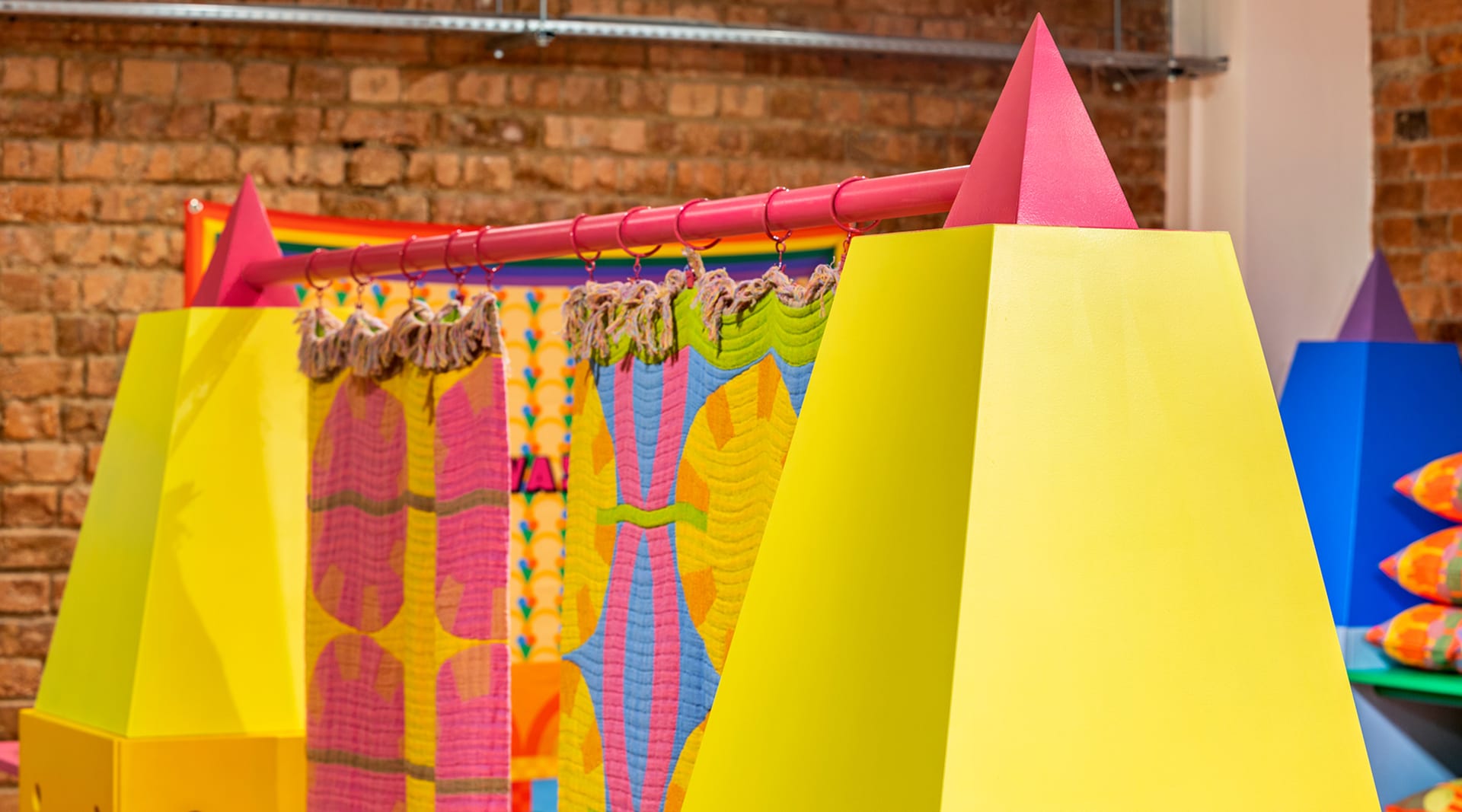 Two yellow pyramid pillars with pink tips hold a colorful tapestry that hangs between them. The image is angled with the right pyramid closer to the viewer.