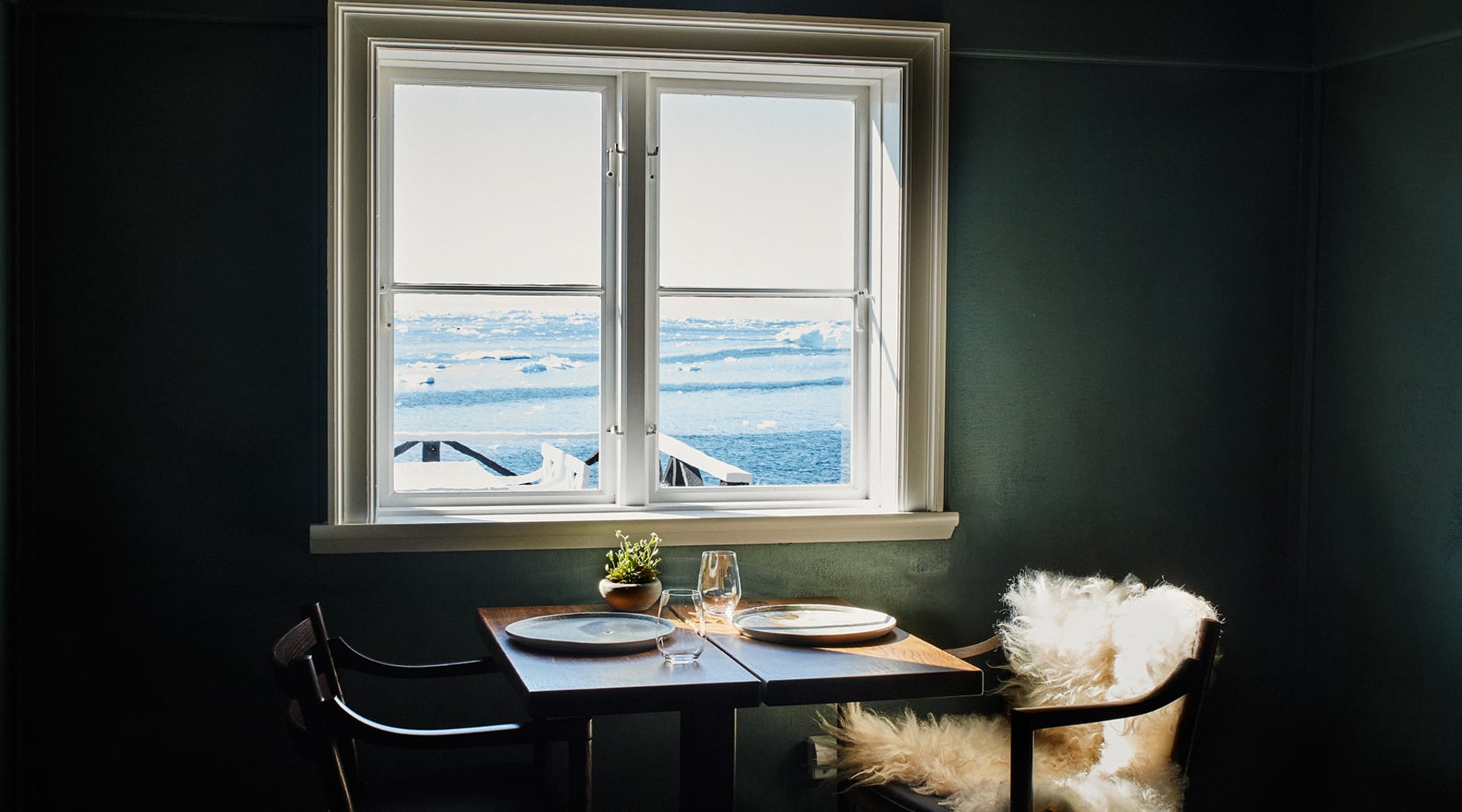 A table is set in front of a window in a room with dark green walls and two chairs. Through the window is an arctic ocean scene.