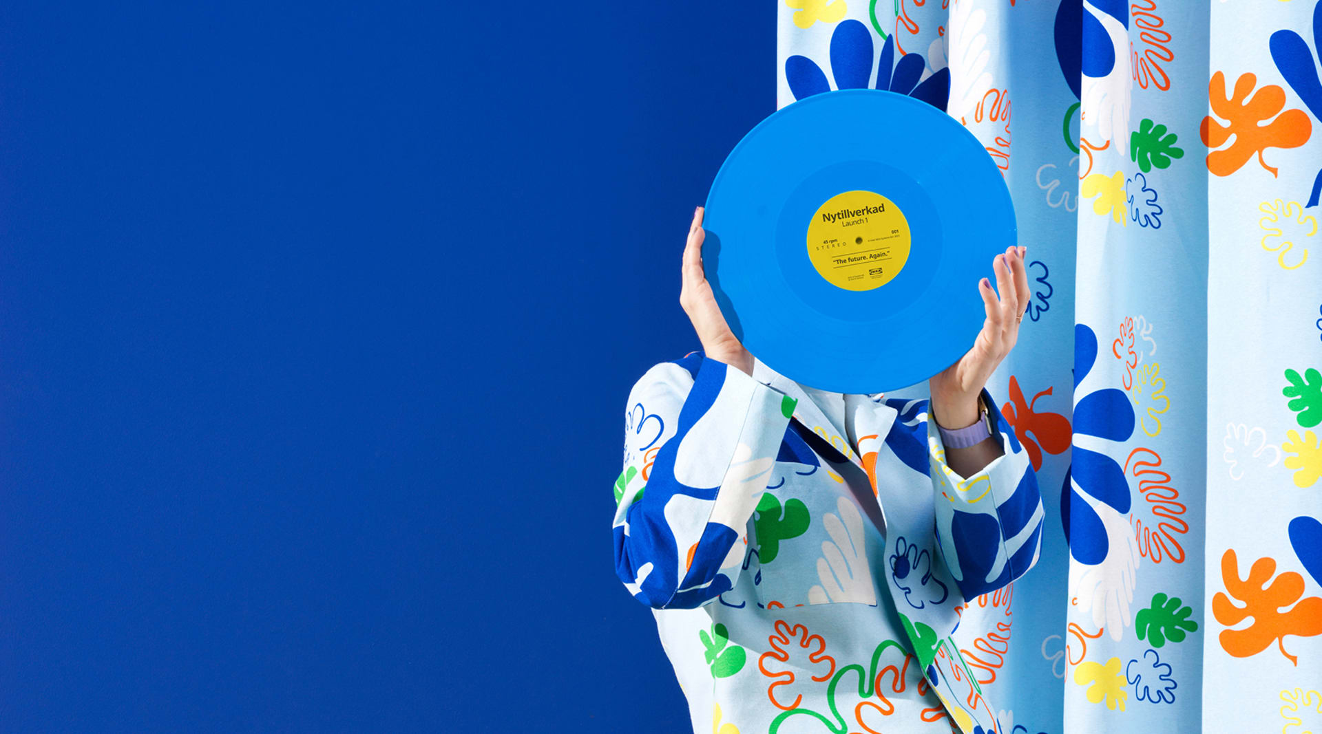 A person stands holding a light blue record in front of their face in a room with a dark blue wall and a blue and orange floral curtain to the right.