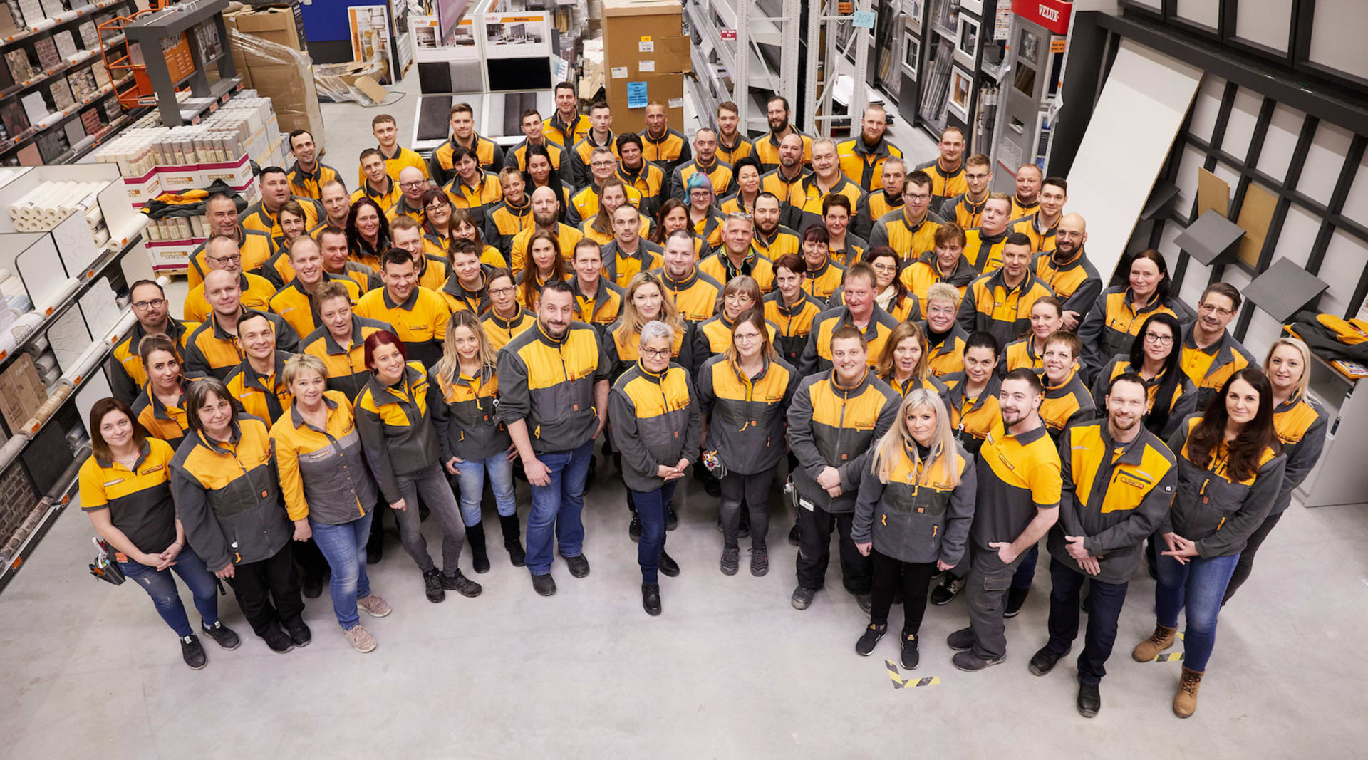 An aerial image taken from ceiling height, of a large group of people smiling up at the camera wearing yellow vests.