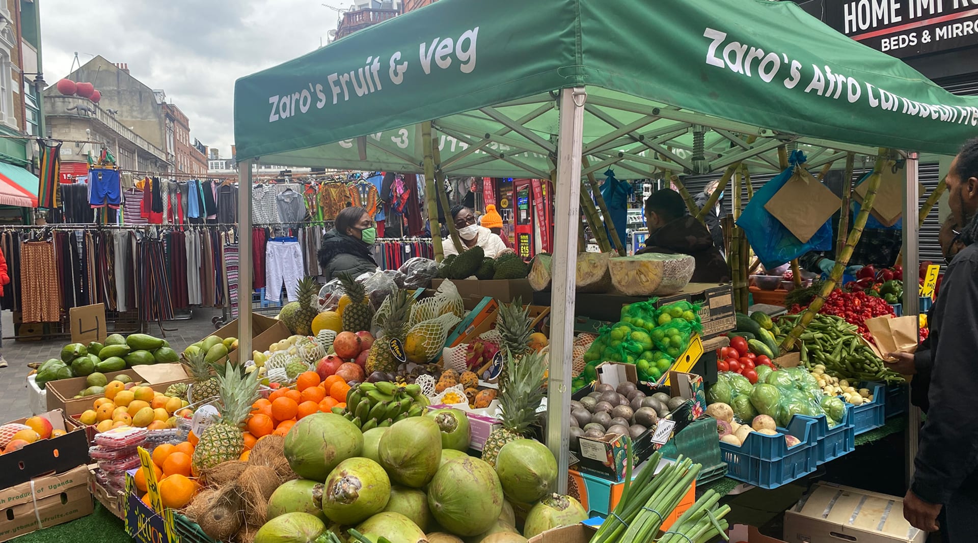 A green awning that reads "Zaro's Fruits and Veg" stands over a large, overflowing fruit stand with three people standing and shopping beneath it in the background.