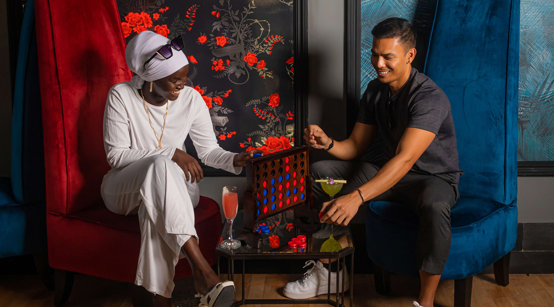 A woman with dark skin wearing a purple headscarf and all white sits across a small circle table from a man with tan skin wearing all black. They play a game on the table, and a cocktail rests on the table in front of each of them.