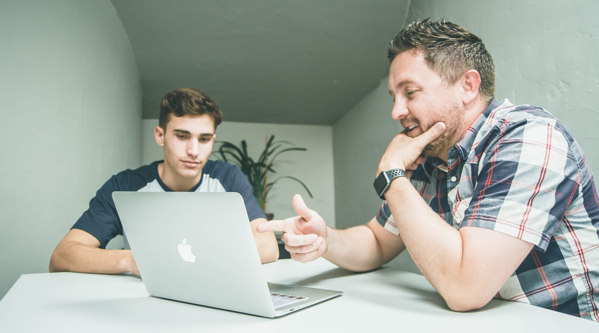 Two white men sit at a table looking at a laptop. The man on the right has a beard and appears older than the man on the left. Both have brown hair.