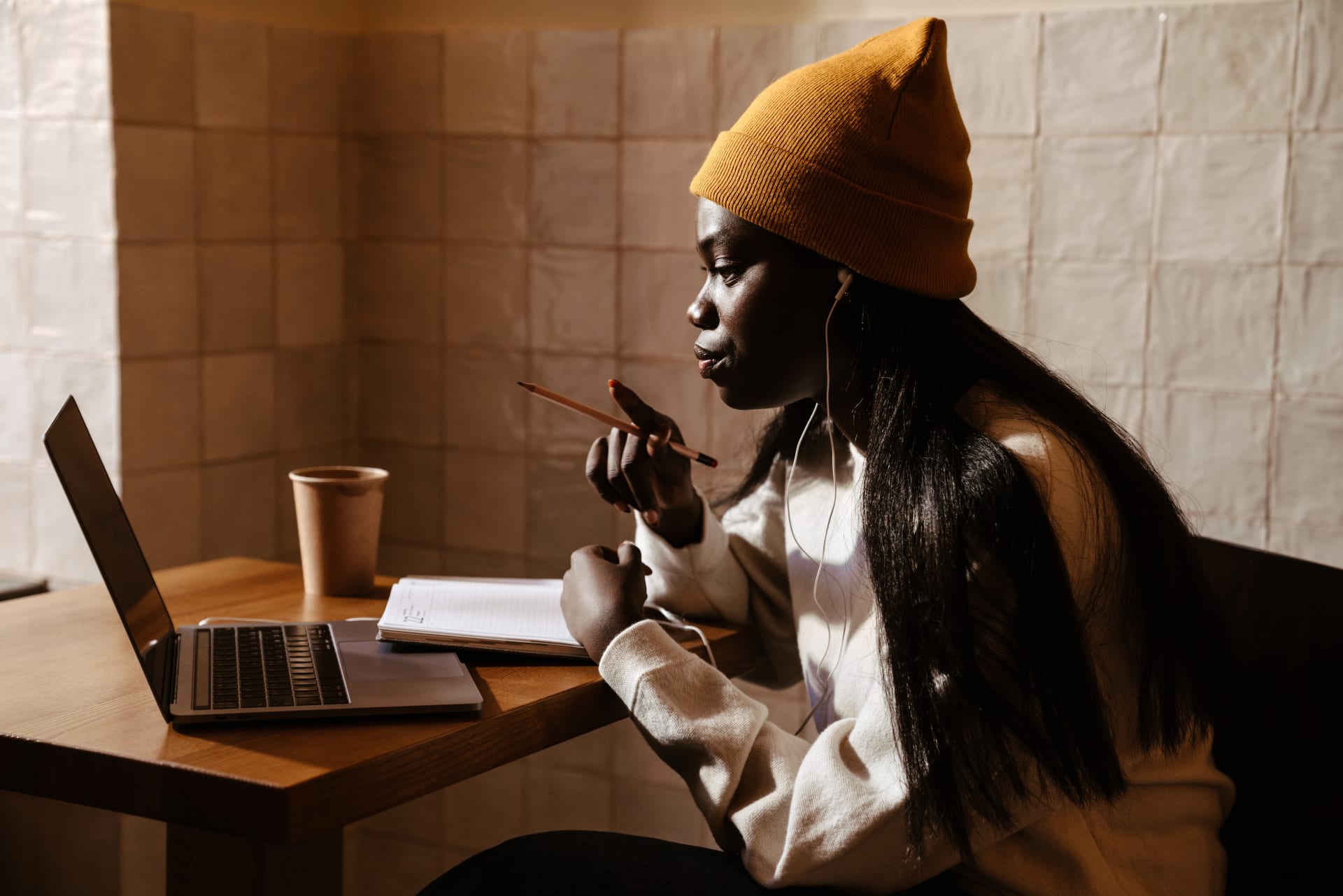 Young black woman in a yellow beanie sat in a cafe using a laptop and wearing wired headphones