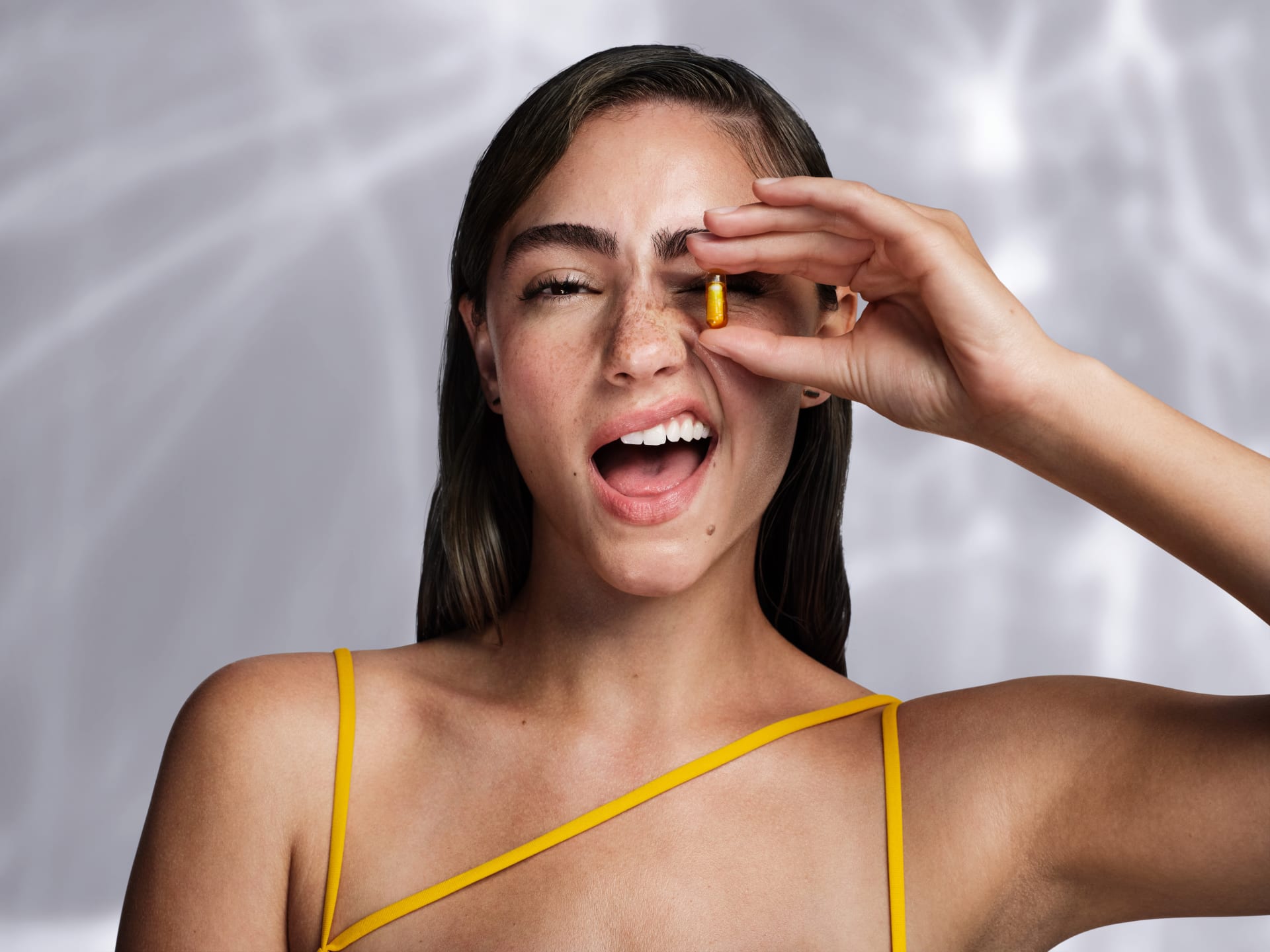 White woman with brown hair and freckles is winking and holding a yellow capsule in front of her left eye She wears a top with thin yellow crisscross strap