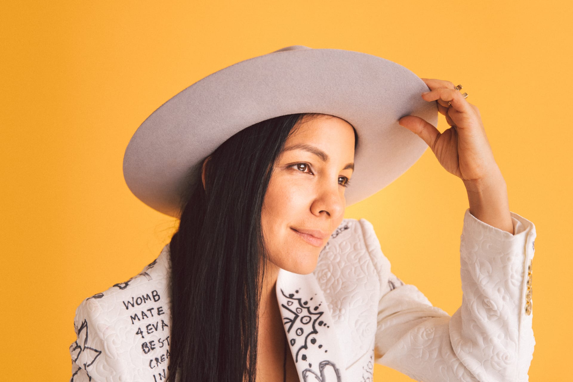 A woman with long dark hair wearing a white jacket covered in graffiti and a grey broad brimmed hat smiles looking off camera