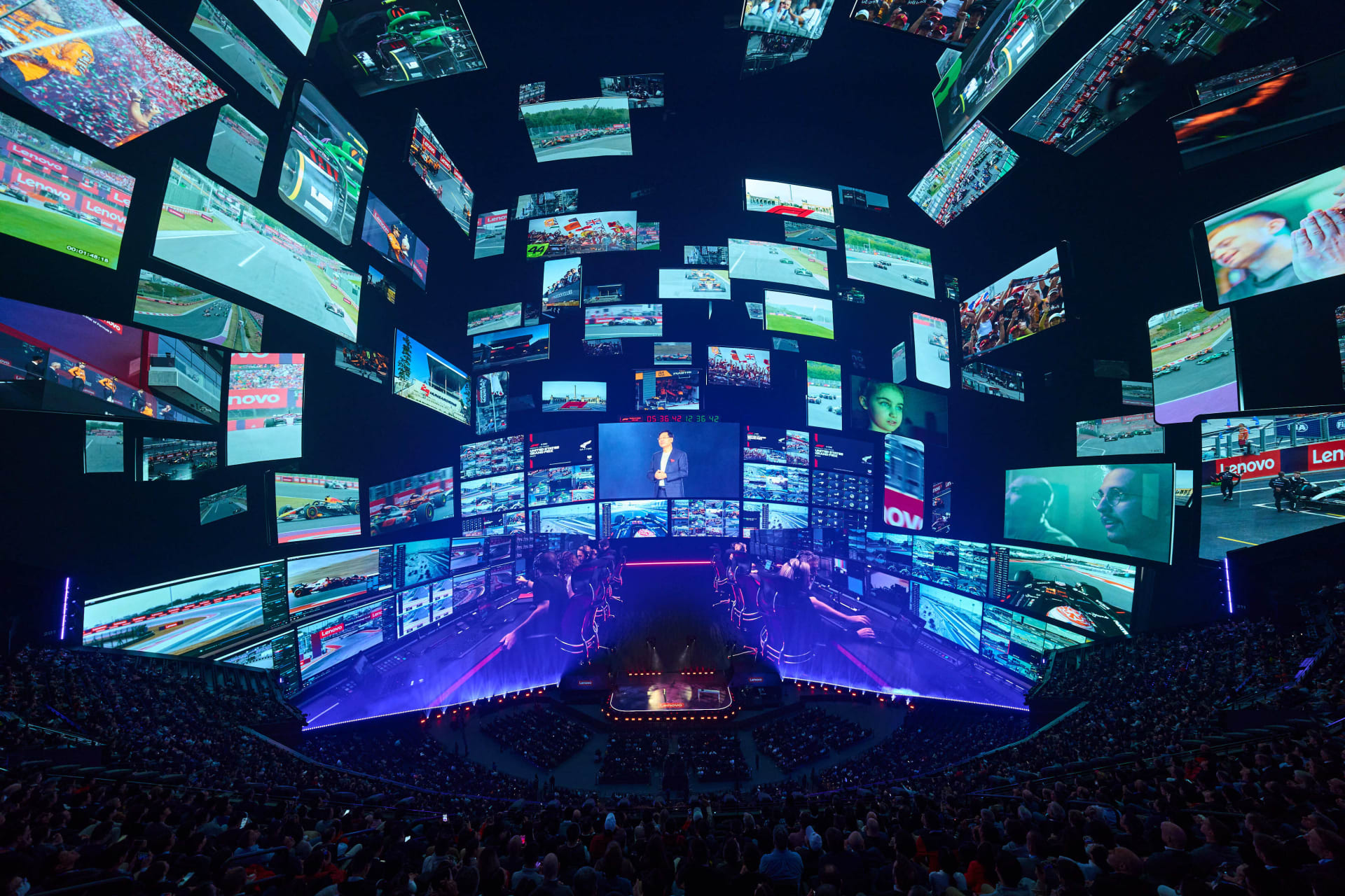 A large audience watches a presenter on stage surrounded by numerous screens displaying AI-driven racing footage at a CES technology event.