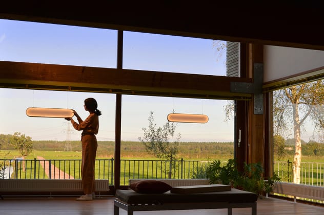 A person in a dimly lit room adjust a pill-shaped light fixture hanging in front of wall-to-ceiling windows as the sun sets in the distance.