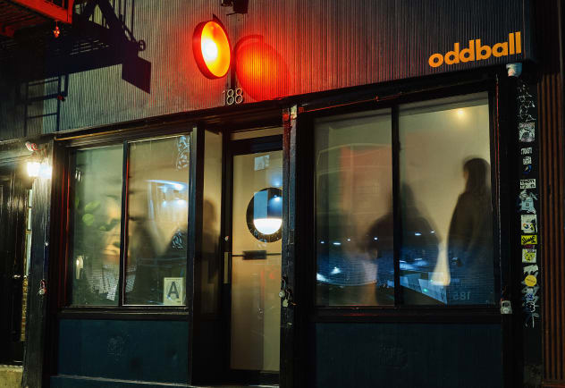 Facade of the Oddball cocktail bar on  a wet paved street by night. There is a black fascia with oddball logo in lower case orange lettering. Patrons are visible through the frosted glass inside. An orange sign casts a glow in the dark.