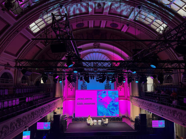 A view of the stage at Shoreditch Town Hall with two people in conversation in front of a pink and blue screen in the distance, with an arched glass ceiling above.