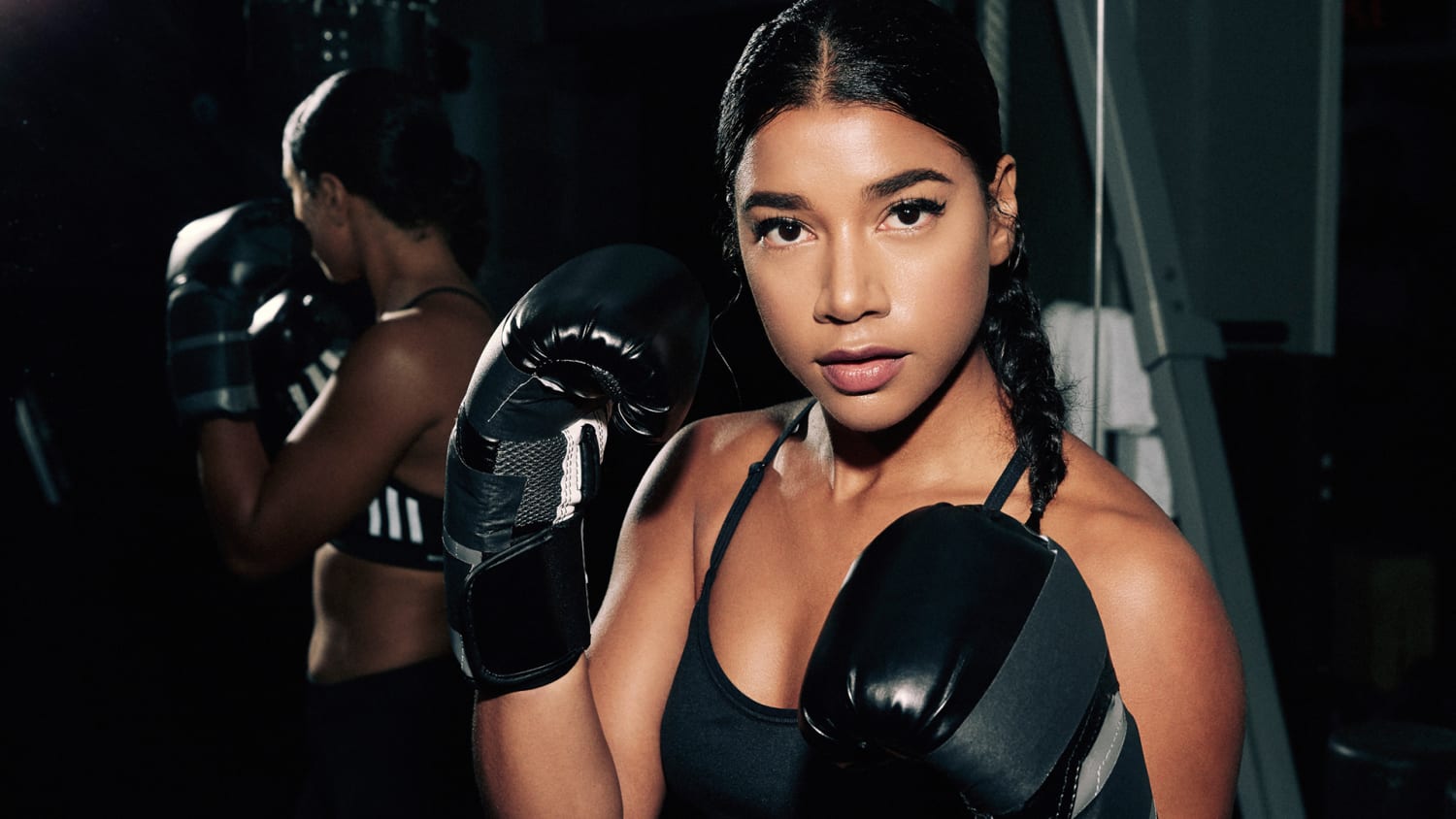 Young woman facing forward in black tank top and boxing gloved. Mirror behind her, reflecting her image.